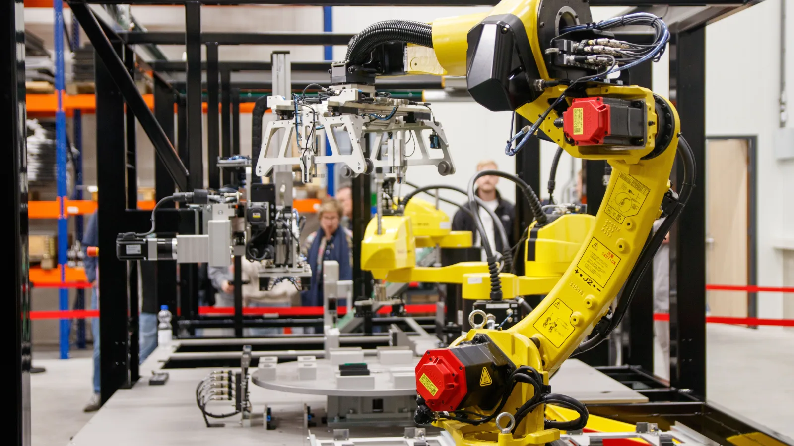Žuta industrijska robotska ruka u proizvodnom pogonu, dok je ljudi promatraju u pozadini.